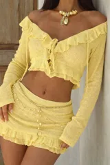 Conjunto de dos piezas color amarillo, compuesto por un top de manga larga con escote off-shoulder, volados y cierre frontal con lazos, y una minifalda a juego con detalle de volado en el ruedo.