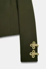 Cazadora militar de color verde oscuro, de cuello subido y manga larga. Cuenta con detalles de alamares y cordones dorados en los hombros y puños, y cierre frontal con botones dorados metálicos.