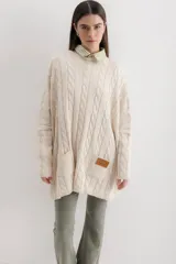 Sweater de punto color camel con diseño de trenzas verticales y acabado brillante. Presenta una silueta holgada tipo poncho, cuello redondo, mangas largas con puños ajustados y bolsillos frontales con detalle de etiqueta de cuero.