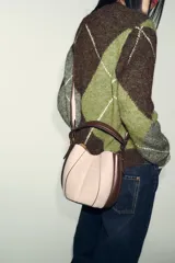 Bolso tipo saca con diseño estructurado, confeccionado en una combinación de tejido de lana en tono beige y laterales de cuero sintético marrón. Presenta costuras decorativas verticales, asa de mano superior y una correa bandolera extraíble con detalle de franjas. Cuenta con cierre mediante botón imantado.