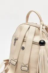 Mochila de símil cuero texturizado color beige, con asa superior, tiras regulables para la espalda y cierre principal metálico. Presenta un detalle trenzado vertical en el frente y un accesorio con flecos y cuenta negra en el lateral.