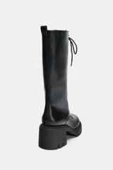 Bota de caña media con cordones frontales, confeccionada en cuero sintético negro. Presenta un tacón ancho en bloque y suela dentada de goma. Cuenta con cierre lateral y tirador en la parte trasera.