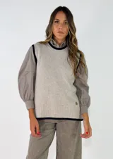 Chaleco de punto en color beige con detalles de ribetes en contraste negro en cuello, sisas y ruedo. Presenta un diseño de corte recto y un pequeño aplique decorativo en la parte inferior.