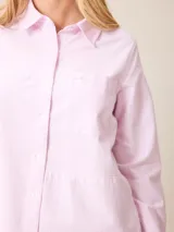 Camisa de manga larga con cuello clásico y cierre frontal de botones, confeccionada en tela de algodón con diseño de rayas verticales finas en color rosado y blanco. Presenta un bolsillo frontal en el pecho con detalle de etiqueta pequeña.