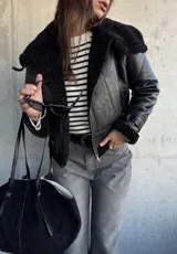 Campera tipo gamulán de cuero sintético negro con interior y detalles de cuello y puños en piel sintética negra. Presenta cierre frontal, correas ajustables en los puños y diseño de corte clásico.