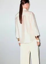 Camisa de satén color crudo con apliques de strass, cuello camisero, manga larga con puños abotonados, diseño recto y cierre frontal de botones ocultos.