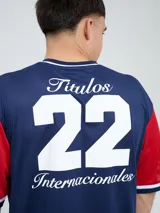 Remera de fútbol Umbro del Club Nacional de Football, de diseño tricolor con cuerpo azul marino, mangas y canesú rojos, y cuello en V con detalles en blanco y azul. Incluye el logo de Umbro en el pecho derecho y el escudo del club en el izquierdo.