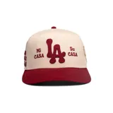 Gorra GQH de visera plana, color blanco con visera bordó. Presenta bordados frontales en relieve con las letras "LA" y el texto "Mi Casa Su Casa" en color bordó.