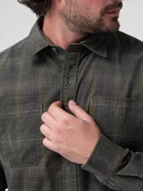 Camisa de manga larga confeccionada en algodón orgánico con textura de cotelé. Presenta un diseño de cuadros escoceses en tonos verdes y grises, cuello clásico, cierre frontal con botones y dos bolsillos frontales con solapa.