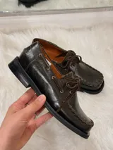 Zapato tipo mocasín de cuero color chocolate, con suela plana de goma negra, cordones al tono y costuras decorativas visibles en el borde superior.