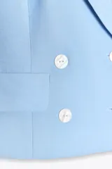 Chaleco estilo blazer sin mangas, color celeste claro, con cuello halter y escote en V profundo. Presenta un cruce frontal con doble botonadura de botones blancos y dos bolsillos de tapeta en la parte inferior.