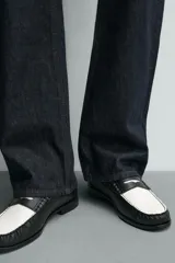 Mocasín de cuero liso, estilo penny loafer, con contraste de color blanco y negro. Presenta un antifaz superior con una abertura central y costuras visibles en el borde. Tiene suela negra de bajo tacón.