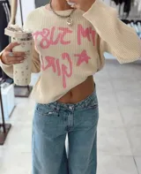 Sweater de punto color beige con diseño de texto en color rosa que dice 'I'm just a girl' y una estrella. Presenta cuello redondo, manga larga y corte holgado.