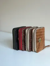 Billetera pequeña de cuero color tostado con textura símil cocodrilo, cierre perimetral de cremallera y detalle de aplique metálico circular en el frente.