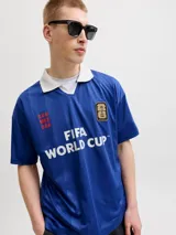 Camiseta de estilo polo con cuello camisero, manga corta y calce relajado. Presenta un diseño en color azul con estampado frontal "FIFA WORLD CUP" y detalles gráficos en contraste. Confeccionada en tejido técnico de poliéster reciclado.