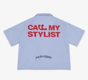 Camisa de corte boxy fit color celeste, con mangas cortas y cuello tipo polo. Presenta un estampado frontal en color rojo con la frase 'CALL MY STYLIST' y un detalle bordado en color negro con la firma 'FACHAPOSTA'.