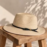 Sombrero tipo fedora de paja color beige, con ala ancha y una tira decorativa negra alrededor de la copa con detalle metálico dorado.
