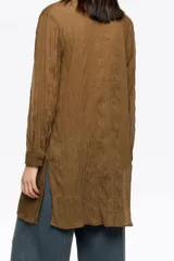 Camisa larga de corte recto, color marrón, confeccionada en tejido plisado. Presenta cuello camisero, botones frontales y mangas largas con puño. Destaca por sus maxilazos colgantes en el escote.