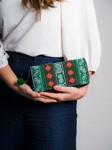Billetera tipo sobre con cierre, confeccionada en tela bordada con motivos tribales en tonos verdes, rojos y blancos. Tiene un tirador con pompón y cuentas de madera.