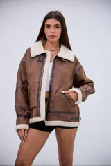 Campera tipo gamulán de corte oversize, confeccionada en cuero sintético marrón con efecto desgastado y forro interno de corderito sintético color crema. Presenta cuello solapa, cierre frontal con cremallera, detalles de tiras con hebillas en el cuello y en los laterales, y costuras reforzadas con bandas de cuero oscuro.