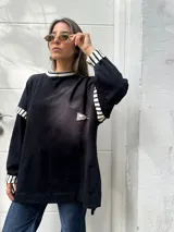 Buzo oversize de color negro con detalles de rayas blancas y negras en el cuello, puños y hombros. Presenta un pequeño parche bordado en el pecho.