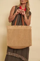 Bolso tipo shopper de rafia trenzada en color tostado, con asas de hombro en cuero sintético marrón y detalle de flecos en el borde superior.