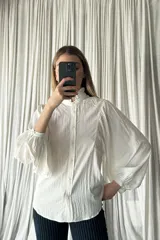 Camisa blanca de textura arrugada con cuello alto tipo mao con volados, cierre frontal de botones y mangas largas abullonadas con puños ajustados.