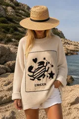 Sweater oversize de tejido de punto en color crudo, con cuello redondo y mangas largas. Cuenta con un parche rectangular al frente con diseño bordado a mano que representa el signo zodiacal de Cáncer, acompañado de un corazón, estrellas y la palabra "CANCER".