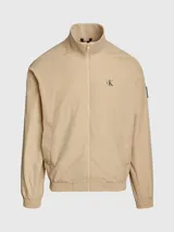 Campera bomber color beige de nailon reciclado, con cuello alto de canalé, puños y bajo elásticos, cierre de cremallera, bolsillos con botón de presión en la cintura, forro de poliéster reciclado e insignia con monograma de Calvin Klein en la manga.