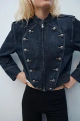 Chaqueta de denim de corte corto, color azul oscuro, con cuello redondo, hombreras y cierre frontal con cremallera. Presenta diseño de inspiración militar con doble botonadura metálica decorativa en el frente, trabillas con botón en los hombros y bajo elástico.