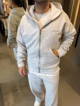 Campera con capucha y cierre frontal, confeccionada en tejido de punto gris jaspeado. Presenta un corte holgado, bolsillos tipo canguro y detalle de logo bordado en el pecho.