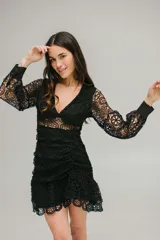 Vestido corto de encaje negro con diseño calado, escote en V, mangas largas con puños y falda con volados. Presenta un corte ajustado con detalle fruncido en la cintura y cierre invisible.