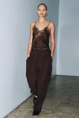 Pantalón marrón de tiro medio y corte carrot wide leg. Presenta trabillas en la cintura, bolsillos laterales ocultos en la costura, pespuntes marcados a tono y una hebilla decorativa en la espalda. Cierre frontal con cremallera, botón y ganchos metálicos.