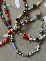 Collar artesanal compuesto por una combinación de cuentas de cristal facetado en tonos gris y bronce, piedras tipo dalmata, cuentas cilíndricas de color crema y cuentas de vidrio azul claro. Presenta un dije central alargado de color azul pastel y terminaciones en cadena de acero.