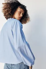 Blusa celeste de popelín, con cuello subido y escote de pico, manga larga abullonada terminada en elástico, y detalles de volantes en el cuello y la botonadura frontal con cierre de botones ocultos.