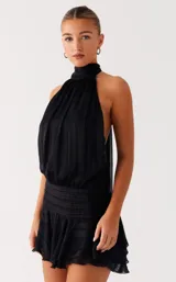 Vestido corto negro con escote halter ajustable con lazo en el cuello y espalda abierta con tira regulable. Presenta cintura fruncida con efecto fajín y falda corta con volados. El tejido es satinado con textura de rayas sutiles.
