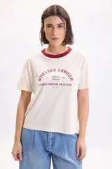 Remera manga corta de color crudo, con cuello redondo en tono bordó y estampa frontal con texto en el pecho.
