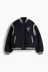 Campera de béisbol azul marino, ligeramente acolchada y confeccionada en tejido polar suave. Presenta cuello, puños y bajo acanalados con rayas blancas, hombros caídos, cierre frontal de botones a presión y bolsillos laterales ribeteados. Lleva un bordado en el pecho con texto que incluye "MILANO FIRENZE ITALIA".