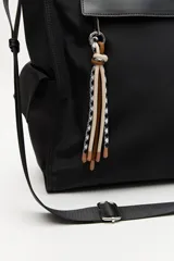 Bolso tipo tote color negro, con doble asa superior y correa desmontable. Presenta un bolsillo frontal con solapa, bolsillos laterales y un detalle colgante decorativo con tiras de colores.
