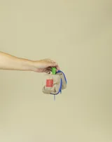 Monedero pequeño de tela color beige con cierre azul y tira de agarre color verde. Incluye un parche rectangular rojo con logo y gancho metálico para colgar.