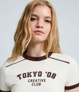 Remera de manga corta color marfil con detalles marrones en contraste en el cuello, costuras de los hombros y puños. Presenta una estampa frontal con la inscripción "TOKYO '08 CREATIVE CLUB" en color marrón, corte ajustado al cuerpo y diseño de estilo urbano.