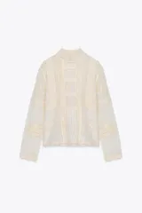 Blusa de manga larga color crema, con cuello alto y detalles de encaje y bordados florales. Presenta paneles verticales con entredoses y un tejido semitransparente.