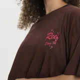 Remera blanca de corte oversized, con cuello redondo y mangas cortas. Presenta un pequeño estampado en el pecho con el logo de la marca Reef en color rojo y detalles de estrellas.