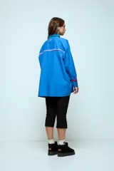 Camisa azul de corte oversize, con cierre frontal de botones y detalles de cintas onduladas en colores contrastantes a lo largo de la botonadura y en los puños. Presenta cuello clásico y mangas largas.