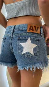 Short de jean azul con ruedo deshilachado, tiro medio y detalle de estrella plateada brillante en el bolsillo trasero. Incluye etiqueta de cuero sintético con logo en la parte posterior.