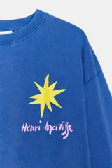 Remera oversize de manga larga color azul, con efecto lavado, cuello redondo y detalle de estampación inspirada en Henri Matisse con una estrella amarilla en el pecho y formas abstractas blancas en la manga.