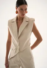 Chaleco sastrero color beige con diseño asimétrico en el bajo, escote en V, botones forrados y hombreras que aportan estructura.