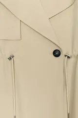 Trench corta de color beige con cuello de solapa y manga larga. Cuenta con bolsillos delanteros, cintura ajustable con cordones, bajo abullonado y cierre frontal cruzado con un botón visible. El interior está forrado con tela a cuadros.