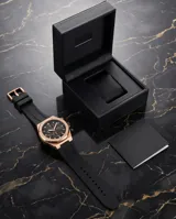 Reloj cronógrafo con caja de aleación metálica en tono oro rosa y esfera negra. Presenta indicadores de hora, subesferas funcionales y correa de caucho negra con cierre de hebilla.