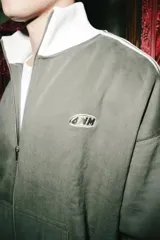Campera deportiva de color verde oliva con cierre frontal completo, cuello alto, puños y ruedo acanalados en color blanco. Presenta franjas laterales blancas en las mangas y bolsillos frontales tipo canguro.